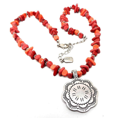 Front view of the retro vintage Chaps pendant necklace. The necklace is beaded with orange and red color nugget style gemstone beads. The pendant, clasp, and chain and darker silver tone in color. The pendant has a flower like scalloped shape to it with a stamped line and circle floral design. There is a lobster claw clasp and extender chain at the end.