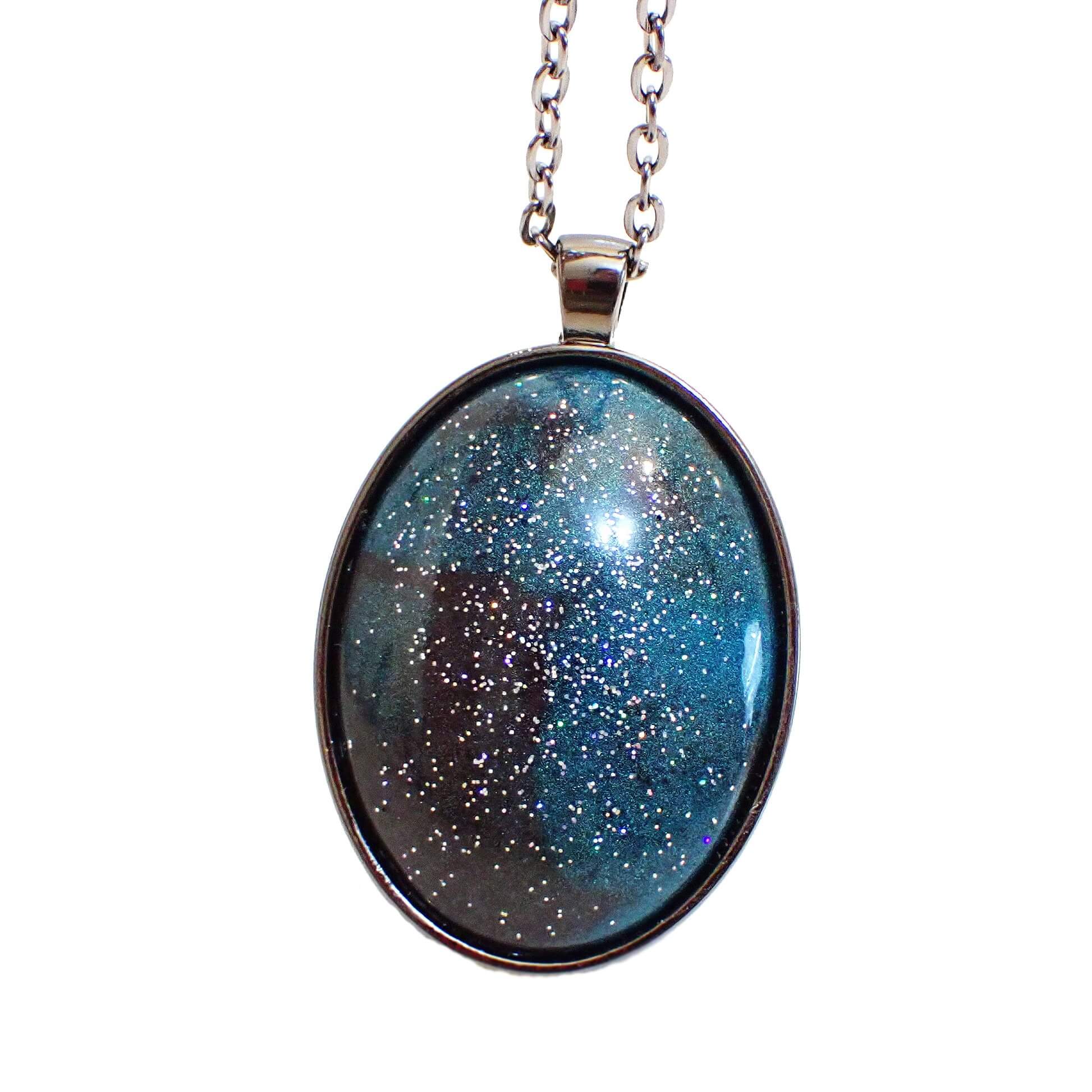 Enlarged front view of the handmade Goth pendant necklace. The metal is gunmetal gray in color on the chain and pendant setting. The pendant is a large domed oval with pearly dark gray and teal blue resin. There is silver holographic glitter embedded in the resin for tiny specks of sparkle and flashes of color as you move around in the light.