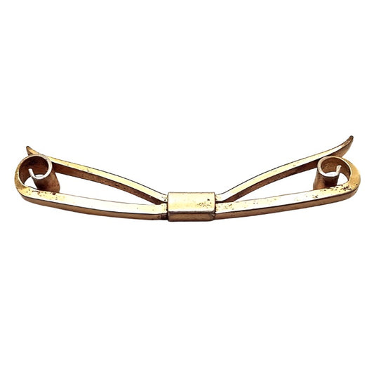 Top view of the 1930's Art Deco vintage collar clip. It is brass metal that is a darker gold tone in color. The front has a bar with curled ends that spiral inward. The back has an angled bar that has slight curves at the ends. Both bars are held together with a rectangular middle area. There are some tiny dark spots on the metal from age.