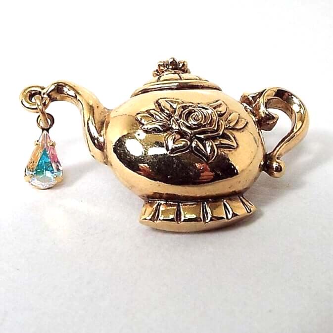 Front view of the Retro vintage Avon teapot tack pin. The metal is gold tone in color. The tea pot is 3D style with a fancy look that has a rose flower in the middle and a teardrop shaped AB rhinestone hanging from the spout. The AB rhinestone is clear with flashes of other colors as you move around. 