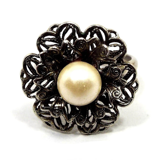 Picture of the top of the adjustable ring. Top has a filigree floral design with two layers of petals. In the middle is a large imitation pearl in an off white color.
