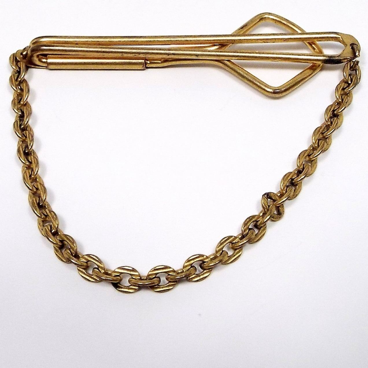 Front view of the 1930's Art Deco vintage Swank tie chain bar. The metal is gold tone in color. There is an open wire style bar that is long and thinner in the front and has a wider angled open area in the back. Rolo chain hangs down from either side of the bar. The links have a textured line pattern on them.