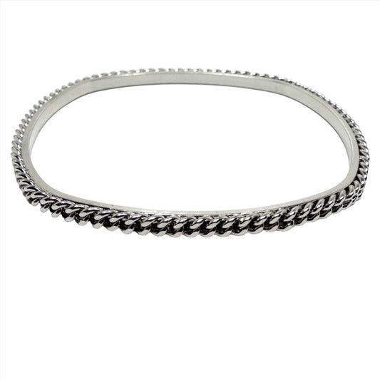 Angled front and side view of the retro vintage bangle bracelet. It is silver tone in color and rounded square in shape. Around the outer edge is a row of  curb chain. The inside is smooth.