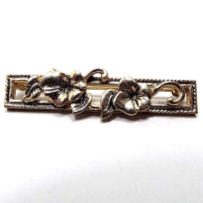 Front view of the retro vintage Avon floral bar brooch pin. It's an antiqued gold tone in color and has an open rectangle with to flowers on the front of it with curled leaves. 