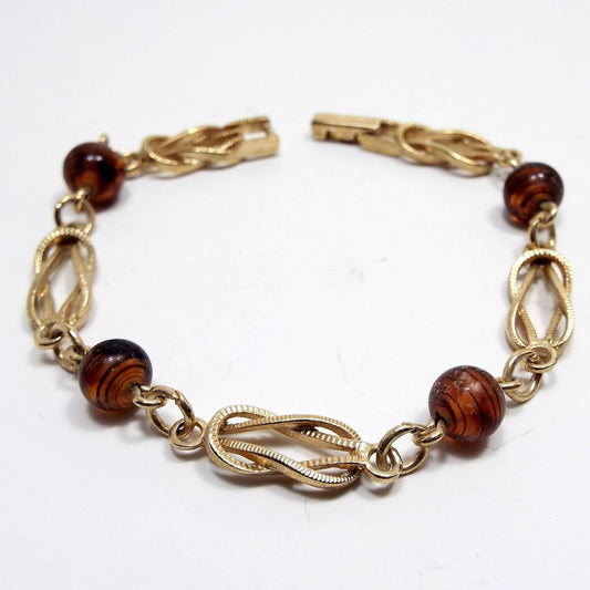 Top view of the retro 1970's Avon vintage beaded bracelet. Metal is gold tone in color. It has alternating links of textured infinity shaped links, round jump rings, and glass faux tortoise beads that are swirled brown and black in color. There is a snap lock clasp on the end.  