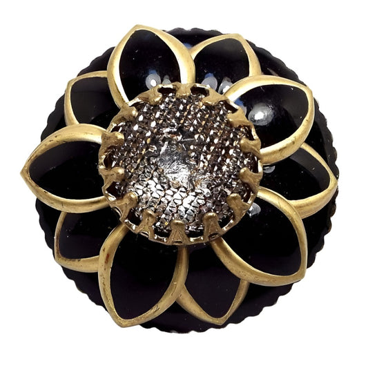 Top view of the large 1990's retro vintage Ollipop floral adjustable statement ring. It's round with a lightly scalloped edge and a sunflower style design. The middle has a textured prong set plastic cab. The petals and background are black enameled with antiqued brass color metal forming the edges of the flower petals. 
