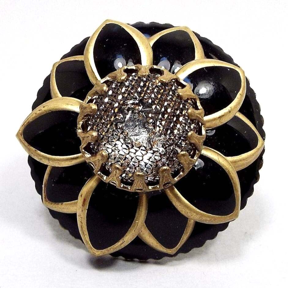 Top view of the large 1990's retro vintage Ollipop floral adjustable statement ring. It's round with a lightly scalloped edge and a sunflower style design. The middle has a textured prong set plastic cab. The petals and background are black enameled with antiqued brass color metal forming the edges of the flower petals. 