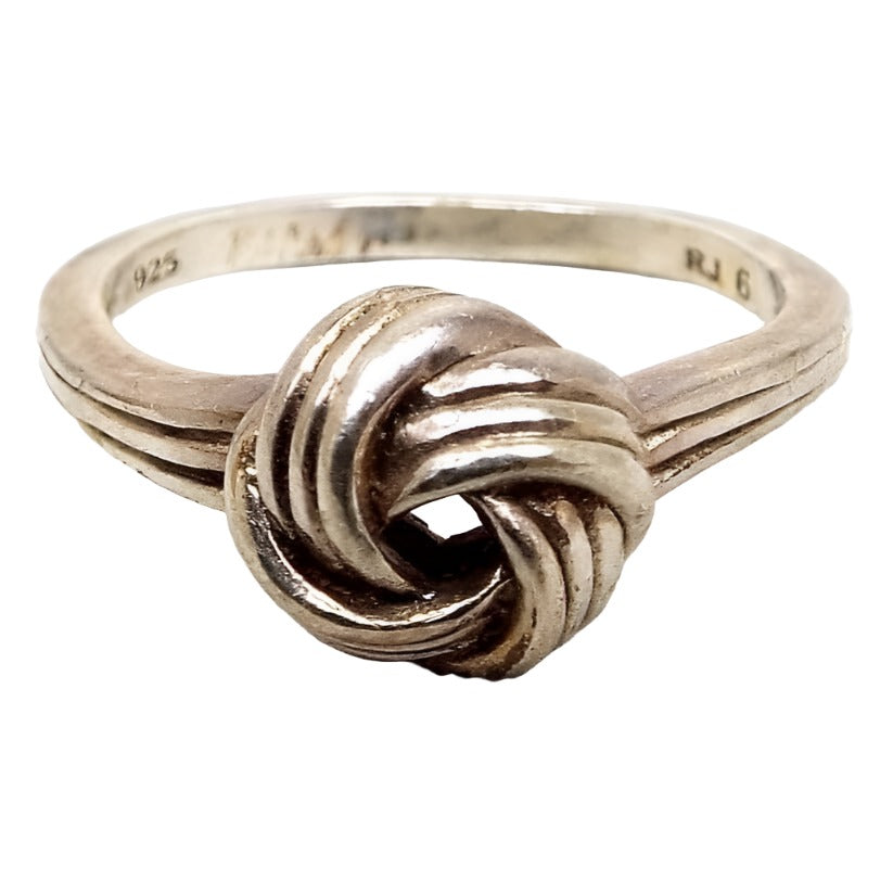Angled front and side view of the retro vintage RJ Graziano sterling silver knot ring. The sterling is slightly darkened from age. There is a knot shape on the top of the ring in the middle. The knot and sides of the band have an indented line design. 925 and RJ 6 can be seen marked on the inside of the band.