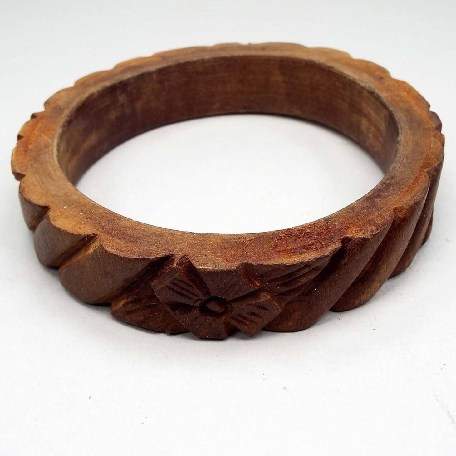 Slightly angled top and side view of the retro vintage wooden bangle bracelet. The wood is dyed brown in color and has rounded carved diagonal stripes all the way around with a carved flower design. The edges and inside of the bangle are flat and smooth.