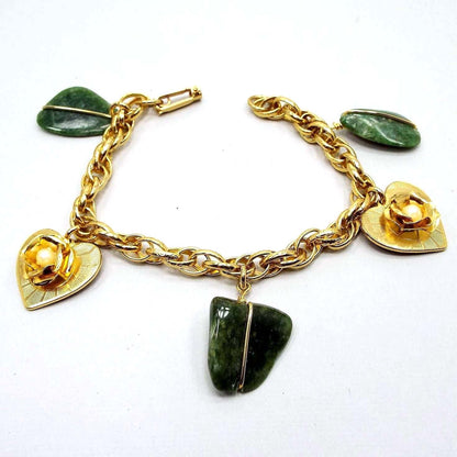 Top view of the retro vintage charm bracelet. The bracelet is gold tone in color and has a textured oval rope chain with a snap lock clasp. The charms alternate between a green flatter style freeform agate gemstone nugget held by a gold tone wire and heart shaped metal charms that have a rose in the middle with plastic faux pearls. 