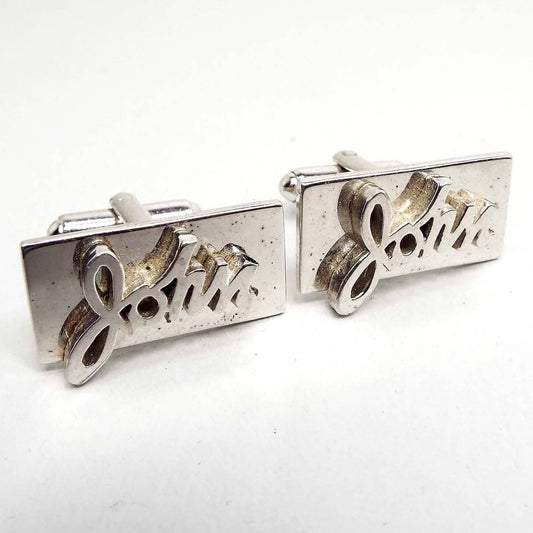 Angled front view of the Mid Century vintage name cufflinks. They are silver tone in color and have the name John in cursive across the fronts on a rectangle background. The levers on the back have rounded levers.