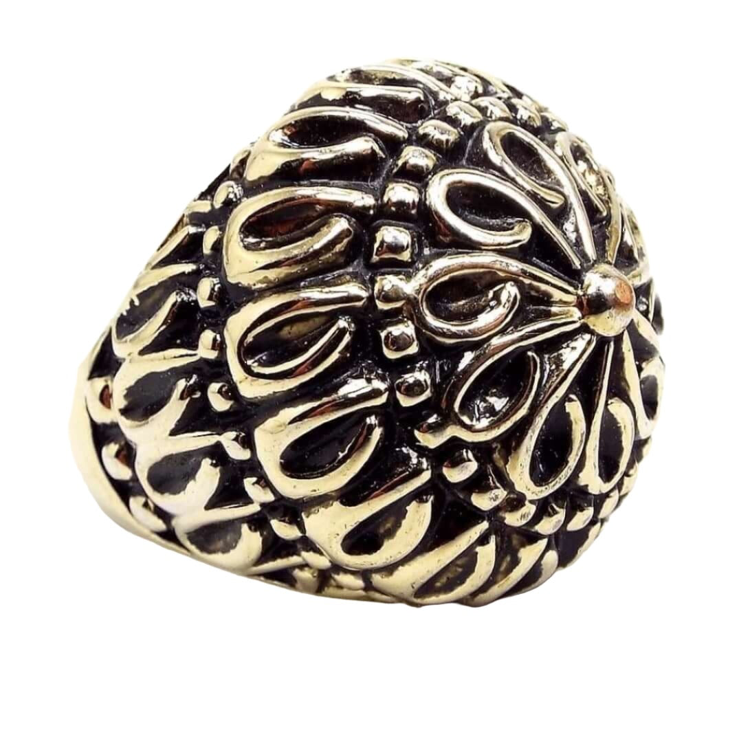 Angled front and side view of the retro vintage dome ring. The plated metal is an antiqued gold tone color with shiny gold color on top of darkened gray areas. The top is rounded with a floral mandala like design. Each petal has a teardrop shaped end. Then there is a row of dots around it, then another row of tear drops and so on until it reaches the bottom band part which is smooth metal. 
