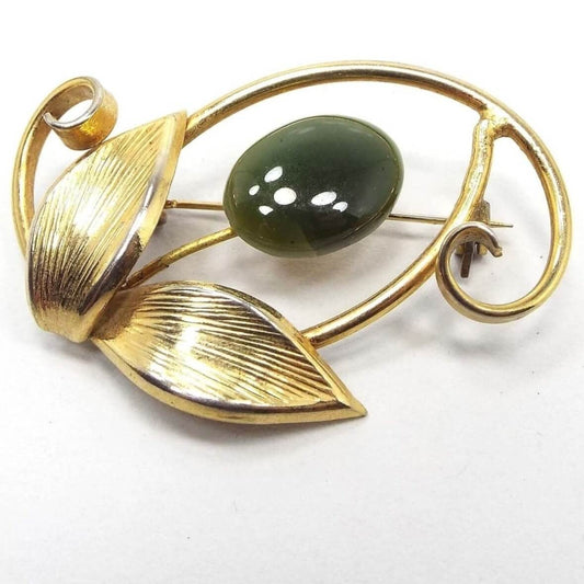 Front view of the retro vintage floral brooch. the metal is gold tone in color. The flower head is a puffy oval green jade gemstone cab. There is a wire stem that goes down to two textured leaves and there are curled areas of metal on each side and around the head of the floor with an open space in between.