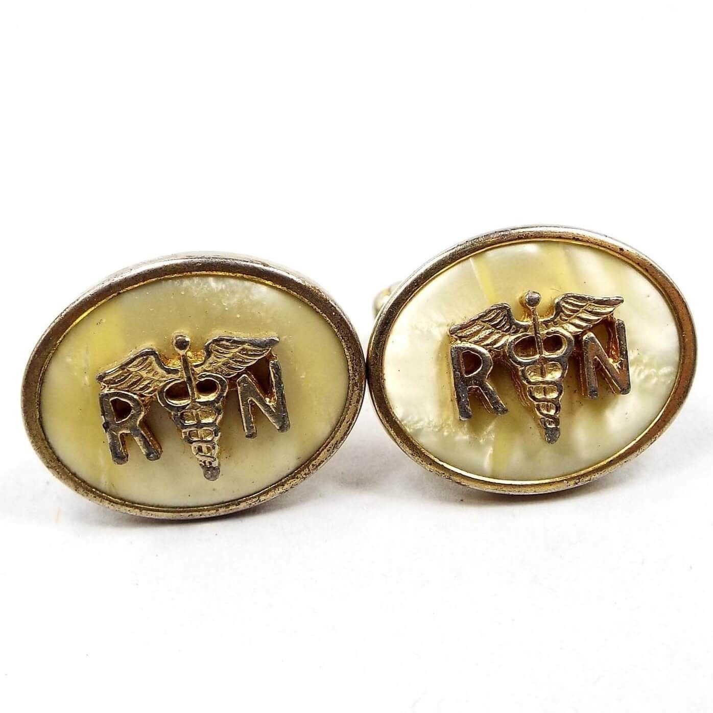 Front view of the Anson Mid Century vintage RN cufflinks. They are oval in shape and the metal is gold tone in color. The inside of the ovals have pearly lucite plastic cabs that are light yellow in color. On top of the lucite cabs is the registered nurse symbol with the letter R and N separated by the Caduceus medical emblem of two snakes entwined around a winged staff. 