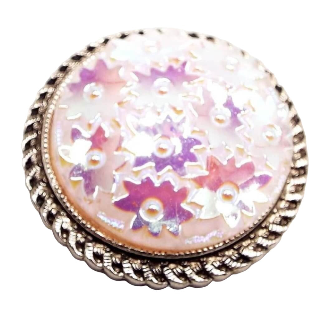 Scarf clip is round with a scalloped edge and gold in color. In the middle is a light pink area covered with small flower shaped larger pieces of glitter that are shimmery purple and pink as you move around. 