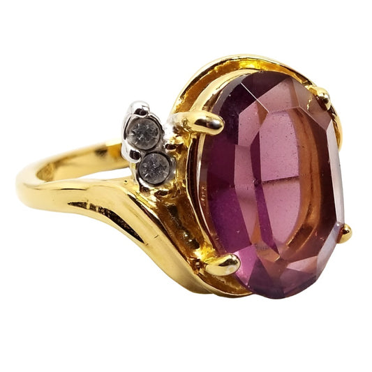 Angled front and side view of the Premier Designs retro vintage rhinestone ring. There is a large prong set chunky faceted oval purple rhinestone in the middle. Each side has two small round prong set clear rhinestones. The metal is gold tone in color.