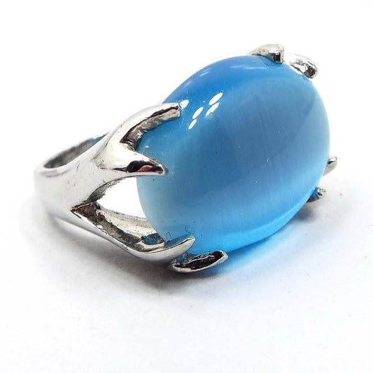Angled front and side view of the retro vintage ring. The top has a large oval glass faux cat's eye cab that is held by a split deer horn or branch style prongs on each side of the ring. There are two prong areas on both sides of the cabochon. It is a lighter blue in color that has a shimmery line area as you move around. The metal is silver tone in color. 