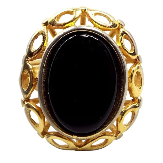 Top view of the retro gold electroplated vintage SA ring. The metal is gold tone in color. The middle has a large flat oval black plastic cab. The outer edge of the ring has a cut out oval design all the way around the cab.