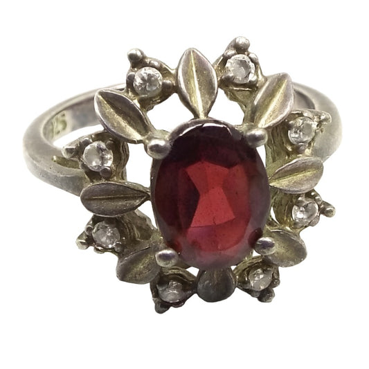 Angled front view of the retro vintage NV sterling silver cubic zirconia and garnet cocktail ring. The top of the ring has a floral design with an oval red garnet gemstone in the middle. There are small silver partially folded petals all the way around the garnet and small round clear CZ stones in between the petals. The sterling is darkened a bit from age for a light silvery gray in color. The 925 marking can be seen on the inside of the band.