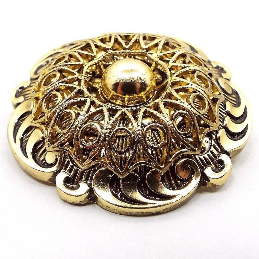 Angled top and side view of the Mid Century vintage scarf clip. It is gold tone in color,. The front part has a plastic filigree domed design with a round ball in the middle. The edges are scalloped with a curled pattern.