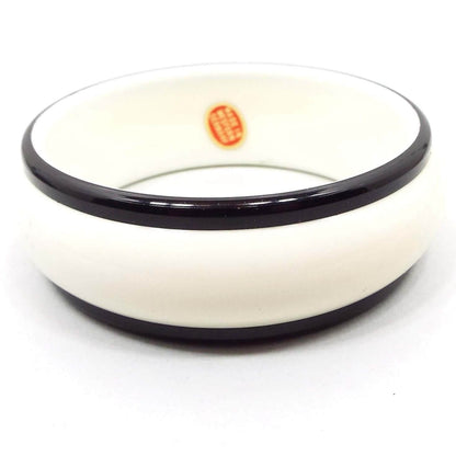 Angled top and side view of the Mid Century vintage plastic bangle bracelet. It is a wider style bangle with a rounded outside edge. The middle part is white plastic and the top and bottom edge are black. An oval red sticker with gold color lettering can be seen on the inside of the bangle that says "Made in Western Germany."