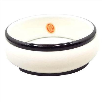Angled top and side view of the Mid Century vintage plastic bangle bracelet. It is a wider style bangle with a rounded outside edge. The middle part is white plastic and the top and bottom edge are black. An oval red sticker with gold color lettering can be seen on the inside of the bangle that says "Made in Western Germany."