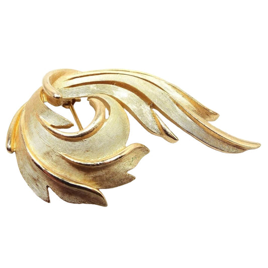 Front view of the retro vintage Crown Trifari feather brooch pin. It's a larger sized brooch with one side curled inward and the other side slightly curved and sloped outward. The inside of the feather design has matte gold tone metal and the edges are shiny gold tone in color. 