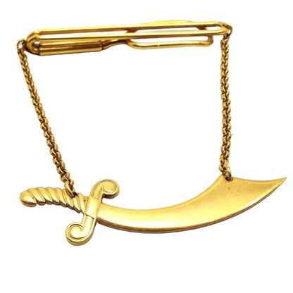 Front view of the 1930's vintage Swank Shriners tie chain bar. The metal is gold tone in color. The top bar is an wire open oval that curves around to the back with another wider open oval on the end. Small rolo chain hangs from either side down to a large scimitar shaped charm.