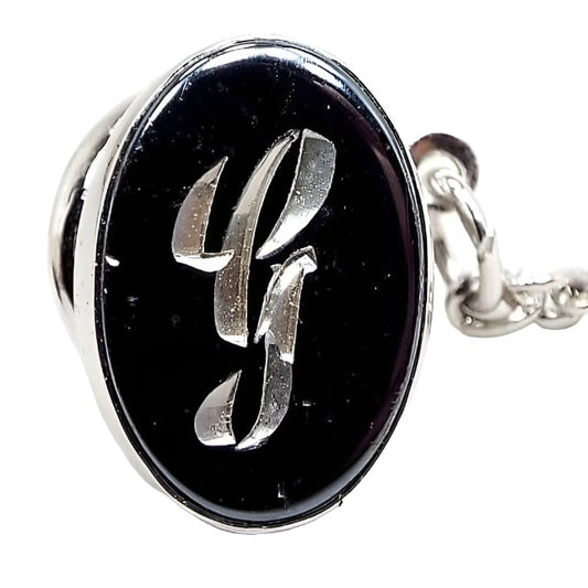 Front view of the Mid Century vintage faux hematite tie tack. The metal is silver tone in color. The tie tack is oval and has a dark metallic gray imitation hemtatite front with a fancy script initial G in the middle. The back clutch has a chain with a bar on the end.