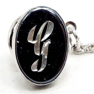 Front view of the Mid Century vintage faux hematite tie tack. The metal is silver tone in color. The tie tack is oval and has a dark metallic gray imitation hemtatite front with a fancy script initial G in the middle. The back clutch has a chain with a bar on the end.