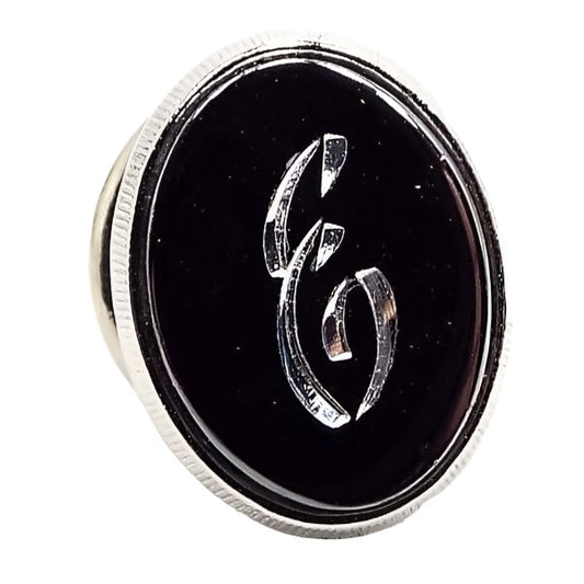 Front view of the Mid Century vintage faux hematite tie tack. The metal is silver tone in color. The tie tack is oval and has a dark metallic gray imitation hemtatite front with a fancy script initial E in the middle. 
