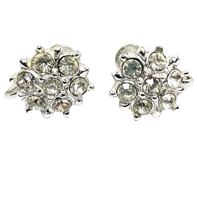 Front view of the 1950's Mid Century vintage rhinestone screw back earrings. The metal is silver tone in color. The fronts have a flower shaped design with round clear rhinestones set around a middle stone. there are small dots of metal in between the petals and middle area. Part of the screws on the back are showing in the photo.