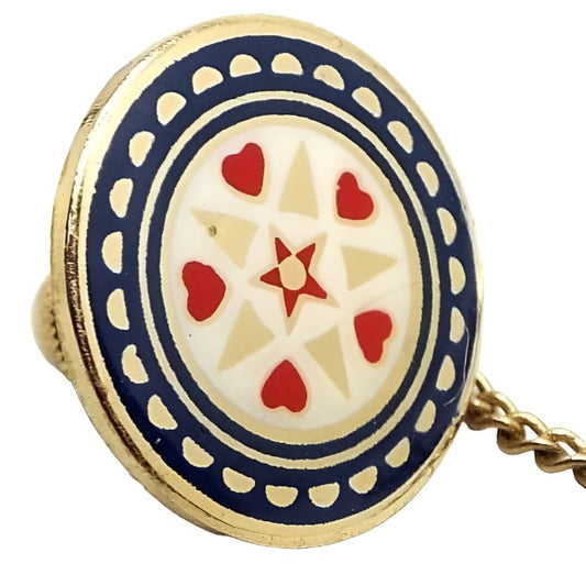 Angled front view of the retro vintage enameled tie tack. The metal is gold tone in color. Tie tack is round and has a Mandala style design with a red star in the middle, a circle row of alternating red hearts and gold color triangles around that with a white enamel background, an enamel blue line, then a wider enamel blue line with gold tone color half moon shapes around the edge. The clutch back has a chain with a small bar on the end.