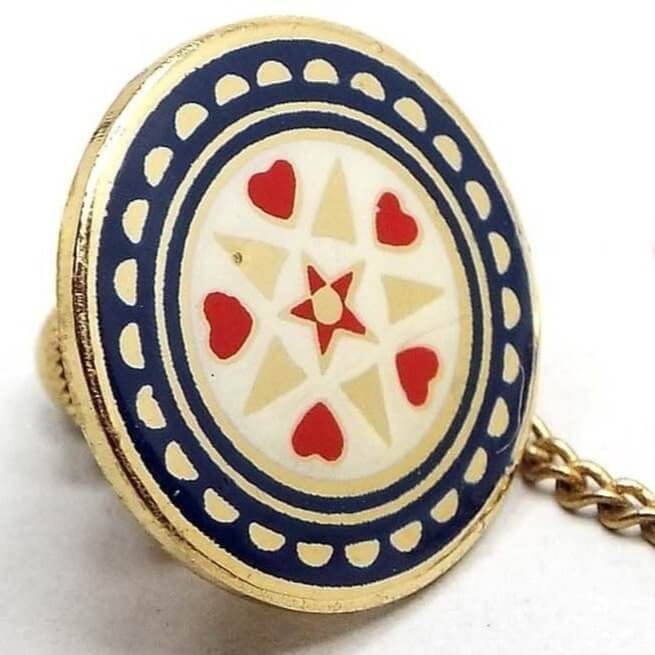 Angled front view of the retro vintage enameled tie tack. The metal is gold tone in color. Tie tack is round and has a Mandala style design with a red star in the middle, a circle row of alternating red hearts and gold color triangles around that with a white enamel background, an enamel blue line, then a wider enamel blue line with gold tone color half moon shapes around the edge. The clutch back has a chain with a small bar on the end.