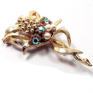 Brooch is gold in color. It looks like a corn stalk with leaves around the stalk on the bottom and curled away from the corn style piece in the middle. Under the corn are AB rhinestones that have flashes of different colors as you move, and under that is a row of imitation pearls. 