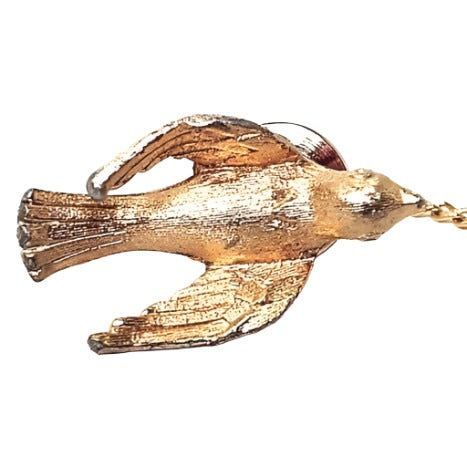 Top view of the retro 1970's Avon tie tack. It is gold tone in color with top view of a 3D dove design. The bird has its wings out and curved towards its tail. 