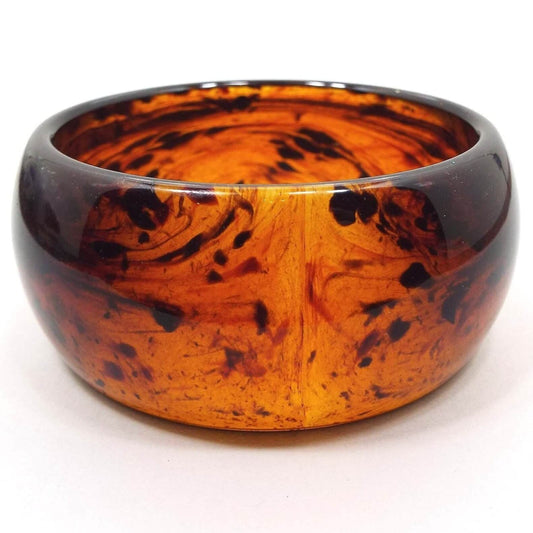 Angled front and side view of the retro vintage lucite plastic bangle bracelet. It is a very wide bangle with curved outer edge. The lucite is semi translucent with areas of orange, brown, and black.