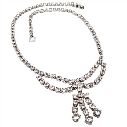 Front view of the 1950's Mid Century vintage rhinestone choker necklace. The necklace has a cup chain design with round prong set clear rhinestones. There is are two looped down areas at the bottom and a chandelier style area with larger rhinestones on the tips. There is a snap lock clasp on the end.