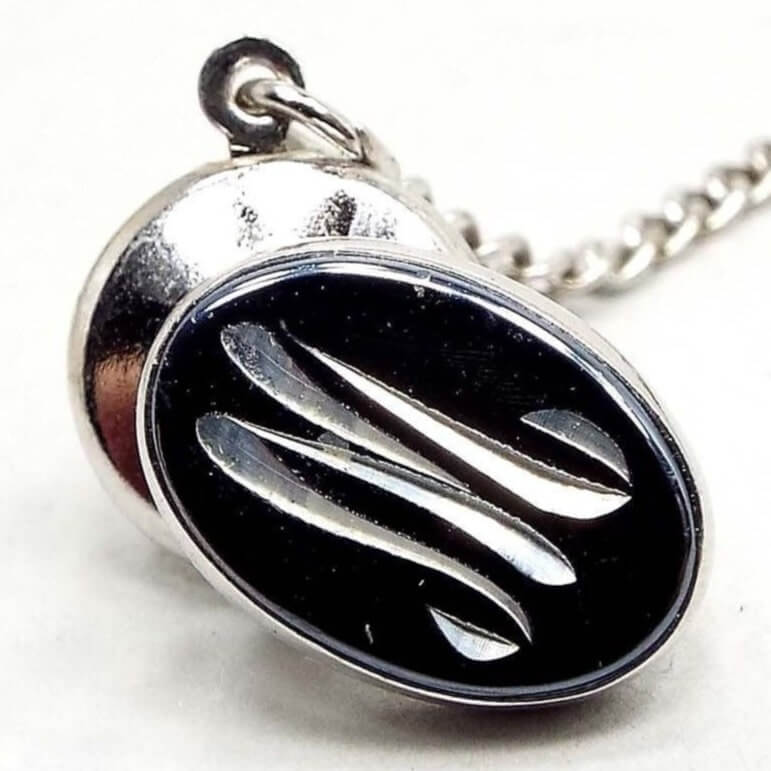 Front view of the Mid Century vintage faux hematite tie tack. The metal is silver tone in color. The tie tack is oval and has a dark metallic gray imitation hemtatite front with a curvy script initial M in the middle. The back clutch has a chain with a bar on the end.