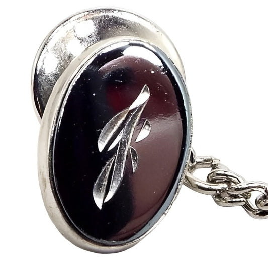 Front view of the Mid Century vintage faux hematite tie tack. The metal is silver tone in color. The tie tack is oval and has a dark metallic gray imitation hemtatite front with a fancy script initial J in the middle. The back clutch has a chain with a bar on the end.