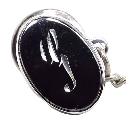 Front view of the Mid Century vintage faux hematite tie tack. The metal is silver tone in color. The tie tack is oval and has a dark metallic gray imitation hemtatite front with a fancy cursive script initial S in the middle. The back clutch has a chain with a bar on the end.