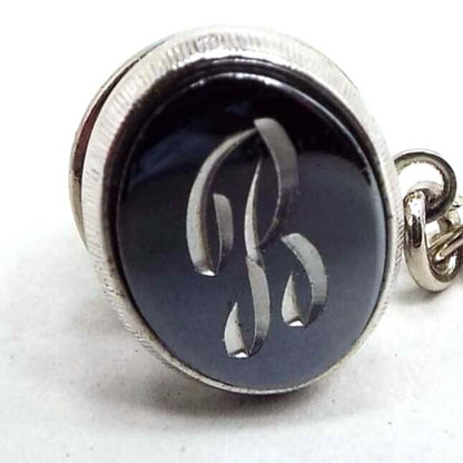 Front view of the Mid Century vintage faux hematite tie tack. The metal is silver tone in color. The tie tack is oval and has a dark metallic gray imitation hemtatite front with a fancy script initial B in the middle. The back clutch has a chain with a bar on the end.