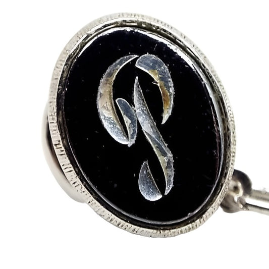 Front view of the Mid Century vintage faux hematite tie tack. The metal is silver tone in color. The tie tack is oval and has a dark metallic gray imitation hemtatite front with a fancy script initial P in the middle. The back clutch has a chain with a bar on the end.