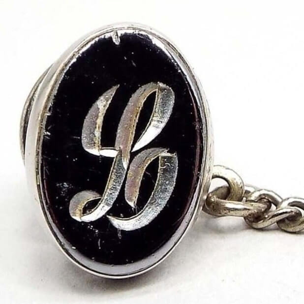Front view of the Mid Century vintage faux hematite tie tack. The metal is silver tone in color. The tie tack is oval and has a dark metallic gray imitation hemtatite front with a fancy script initial L in the middle. The back clutch has a chain with a bar on the end. The bar has some chips from the silver plating showing brass underneath.