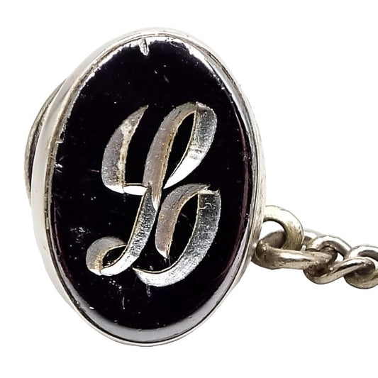Front view of the Mid Century vintage faux hematite tie tack. The metal is silver tone in color. The tie tack is oval and has a dark metallic gray imitation hemtatite front with a fancy script initial L in the middle. The back clutch has a chain with a bar on the end. The bar has some chips from the silver plating showing brass underneath.