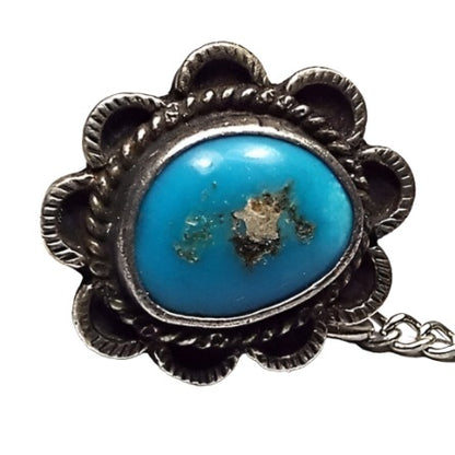 Front view of the retro vintage turquoise tie tack. There is a puffy freeform bezel set oval turquoise cab in the middle. It is blue with an area of textured gray and white stone in the middle. There is a braided rope style edge around the bezel and textured scalloped edges all the way around for a Southwestern style design. There is a chain on the back clutch with a small bar on the end.