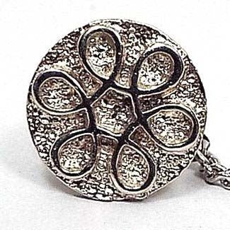 Top view of the retro vintage tie tack. It is round with a textured matte silver tone color front. There is a shiny silver tone color metal spiraled flower design in the middle. The back clutch has a chain with a small bar on the end.