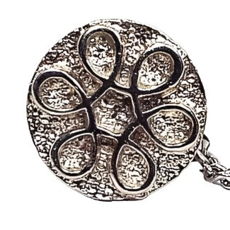 Top view of the retro vintage tie tack. It is round with a textured matte silver tone color front. There is a shiny silver tone color metal spiraled flower design in the middle. The back clutch has a chain with a small bar on the end.