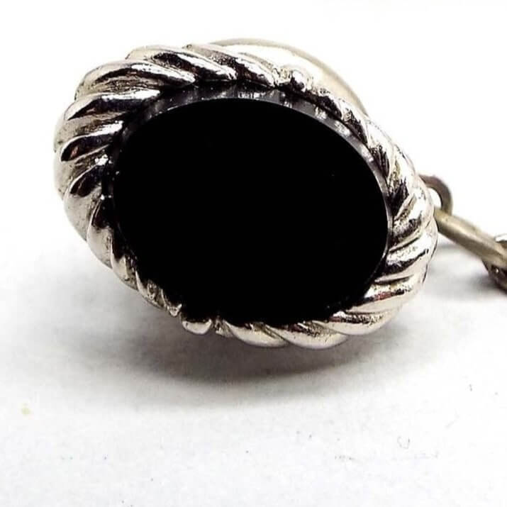 Front view of the Mid Century vintage tie tack. It is silver tone in color with an oval shape. There is a flat black glass cab in the middle with a twisted metal design around the edge. Part of the pin back clutch can be seen on the back that has a chain with a bar on the end.