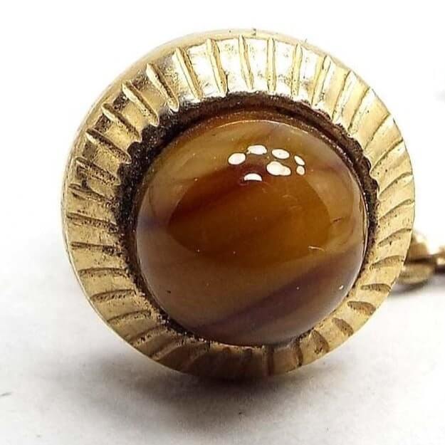 Front view of the Mid Century vintage tie tack. The metal is gold tone in color. It has a round shape with textured lines all the way around the edge. The middle area has a rounded glass cab that has marbled brown and yellow tones. The back clutch has a chain with a small bar on the end.